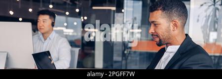 Schöner afrikanischer Geschäftsmann, der mit einem Laptop arbeitet, während er im modernen Büro auf dem Hintergrund eines Kollegen sitzt Stockfoto