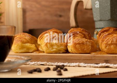 Croissants mit Gelee auf Holzbrett im Stillleben. Kaffeebohnen, eine Tasse Kaffee und eine Kaffeekanne. Holzhintergrund. Stockfoto