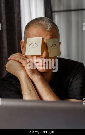 Ein müder Geschäftsmann mit Aufklebern im Gesicht sitzt an einem Tisch im Büro und schläft, die Augen werden auf die Aufkleber gezeichnet. Stockfoto