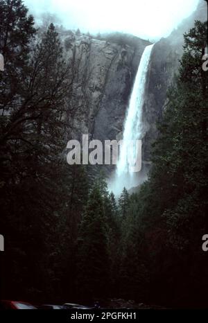 Yosemite-Nationalpark. CA. USA 1984. Yosemite Upper, Middle und Lower Falls. Halten Sie Ausschau nach dem Eiszapfen am Fuß des oberen Wasserfalls im Winter und nach dem brüllenden Abfluss von April bis Juni. Die Yosemite Falls, einer der höchsten der Welt, bestehen aus drei separaten Wasserfällen: Dem oberen Yosemite-Fall (1.430 Fuß), den mittleren Kaskaden (675 Fuß) und dem unteren Yosemite-Fall (320 Fuß). Die beste Aussicht auf die Yosemite Falls bietet die Gegend um Sentinel Meadow und die Yosemite Chapel. Nevada Falls, Bridalveil Falls und Vernal Falls vom Boden des Yosemite Valley aus gesehen. Stockfoto