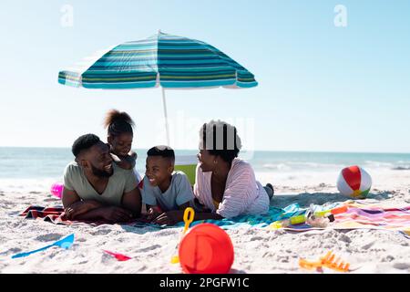 Glückliche afroamerikanische Eltern und Kinder, die sich unterhalten, während sie auf Handtüchern am Sandstrand unter dem Himmel liegen. Kopierraum, unverändert, Familie, zusammen, Sonnenschirm,