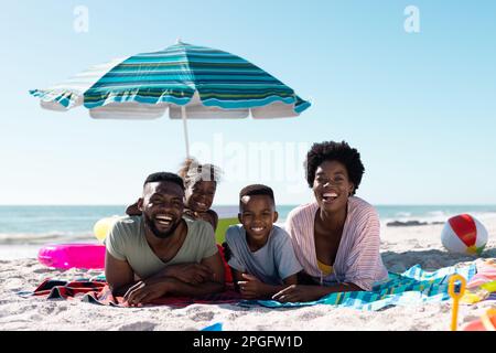 Lächelnde afroamerikanische Eltern und Kinder liegen auf Handtüchern am Sandstrand unter klarem Himmel. Kopierraum, unverändert, Familie, zusammen, Sonnenschirm, Picknick