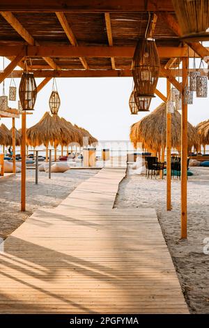 Holzponton am Strand, der zum Meer führt, zwischen Strohschirmen und Strandmatratzen. Bambuslaternen werden vom Strohdach aufgehängt Stockfoto