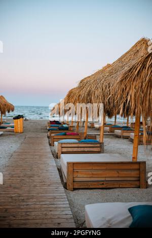 Betten, Matratzen und Strandkissen unter Strohschirmen am Strand. Der Holzsteg führt zwischen den Liegen zum Strand Stockfoto