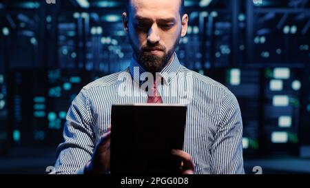 IT-Webtechniker, der im Rechenzentrum mit einem Tablet arbeitet und sich die Rackschränke im digitalen Serverraum ansieht. Professioneller Techniker aktiviert das Hardware-Speichersystem. Handgeführte Aufnahme. Stockfoto