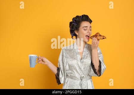 Junge Frau im Seidenbademantel mit Lockenwicklern frühstückt auf orangefarbenem Hintergrund Stockfoto