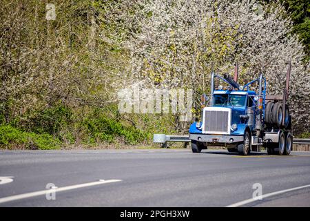 Leistungsstarker industrieller Sattelschlepper mit Motorhaube, klassisch, blauer Sattelzugmaschine mit Day Cab, der einen leeren Sattelanhänger transportiert, um Baumstämme auf der Straße zu transportieren Stockfoto