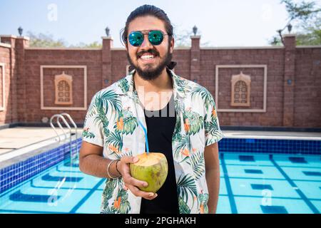 Junger indischer Mann mit Sonnenbrille, der an heißen, sonnigen Tagen im Freien, in den Sommerferien und im Urlaub Kokoswasser genießt. Stockfoto