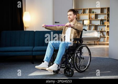 Junger behinderter Mann im Rollstuhl, der zu Hause Übungen mit Gummiband macht. Ein kaukasischer Behinderter, der im Wohnzimmer trainiert. Stockfoto