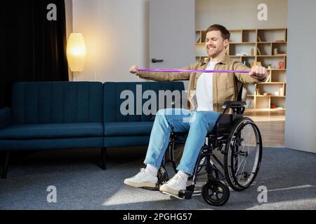 Junger behinderter Mann im Rollstuhl, der zu Hause Übungen mit Gummiband macht. Ein kaukasischer Behinderter, der im Wohnzimmer trainiert. Stockfoto