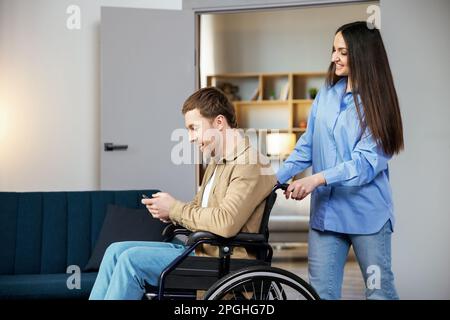 Ein süßer junger Mann im Rollstuhl hält ein Handy, während seine Freundin ihm hilft, sich in der Wohnung zu bewegen. Stockfoto