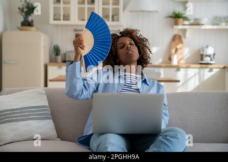 Müde gemischte Frauen, die mit dem Ventilator in der Hand winken, leiden an Hitze, während sie mit einem Laptop auf der Couch zu Hause arbeiten Stockfoto