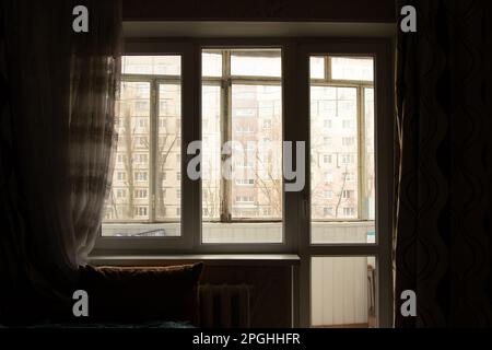 Wohnzimmer in einem Apartment mit Blick auf ein altes Fenster in einem dunklen Raum und ein mehrstöckiges Gebäude außerhalb des Fensters in der Ukraine Stockfoto