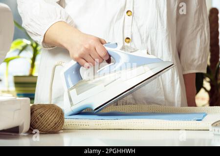 Gekürzte Frau in weißem Hemd bügeln, mit Bügeleisen Dampfgarer für blaufaltige Textilien auf dem Tisch. Schneiderei bereitet Stoff vor, Fertigungsarbeit. Machen Stockfoto