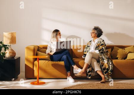 Unternehmerinnen, die sich unterhalten, während sie in einer Bürolobby zusammenarbeiten. Zwei verschiedene Geschäftsfrauen, die auf einer Couch sitzen, in einem Frauenbesitz Stockfoto