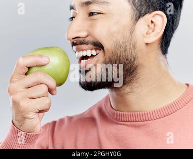 Genießt, glücklich und ein asiatischer Mann isst einen Apfel isoliert auf weißem Hintergrund in einem Studio. Lächeln, Essen und ein Chinese, der von einer Frucht beißt Stockfoto