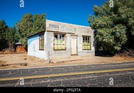 Das geschlossene Postamt, gesehen von der gegenüberliegenden Straßenseite, mit rissigem Pflaster und einem kleinen braunen Schuppen dahinter, im Mojave National Preserve, Kalifornien. Stockfoto