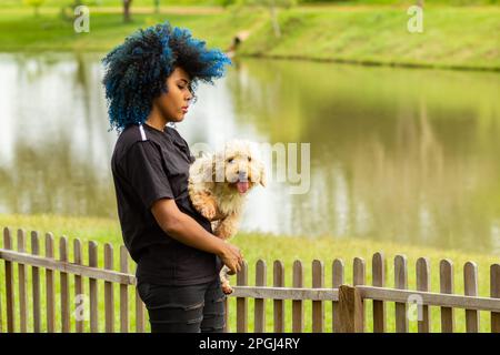 Goiania, Goias, Brasilien – 20. März 2023: Eine junge schwarze Frau mit gelocktem, blau gefärbtem Haar, die ihren Hund auf dem Schoß hält. Stockfoto