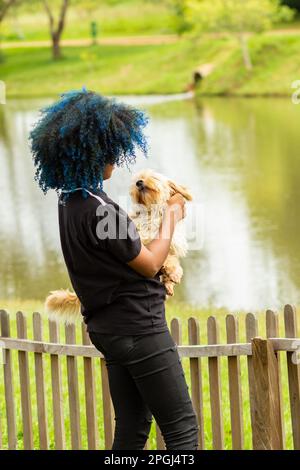 Goiania, Goias, Brasilien – 20. März 2023: Eine junge schwarze Frau mit gelocktem, blau gefärbtem Haar, die ihren Hund auf dem Schoß hält. Stockfoto