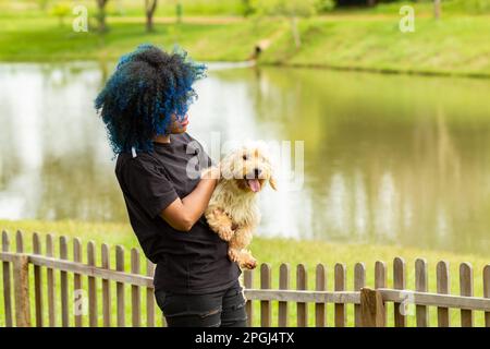 Goiania, Goias, Brasilien – 20. März 2023: Eine junge schwarze Frau mit gelocktem, blau gefärbtem Haar, die ihren Hund auf dem Schoß hält. Stockfoto