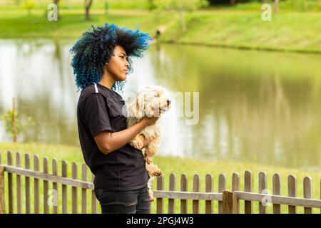 Goiania, Goias, Brasilien – 20. März 2023: Eine junge schwarze Frau mit gelocktem, blau gefärbtem Haar, die ihren Hund auf dem Schoß hält. Stockfoto