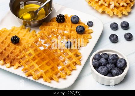 Waffeln und Beeren, ein Löffel in einer Metallschüssel mit Honig, auf einem weißen Teller. Blaubeeren in einer weißen Schüssel. Weißer Hintergrund. Stockfoto