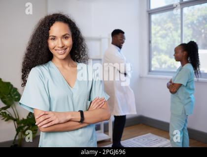 Porträt einer Krankenschwester mit gefalteten, lächelnden, lockigen Armen - mit Team im Hintergrund Stockfoto