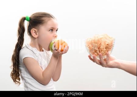 Große Schüssel mit Chips und Obst, wunderschönes kleines Mädchen isst einen Apfel, Kind bekommt eine Schüssel Chips, weißen Hintergrund und Kopierraum, gesund und Junk für Stockfoto