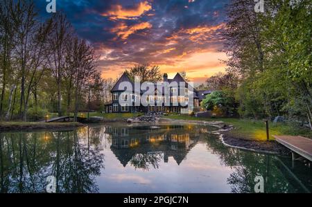 Ein malerisches großes Haus am Ufer eines ruhigen Sees, umgeben von üppigem Grün, das bei Sonnenuntergang eingefangen wurde Stockfoto