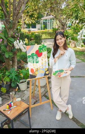 Künstlerkonzept, weibliche Künstlerin, die glücklich ist, Kunst auf Leinwand im Garten zu malen. Stockfoto