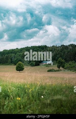 Ein vertikales Bild des bewölkten Himmels über den bewaldeten Hügeln und ein Haus auf dem Feld Stockfoto