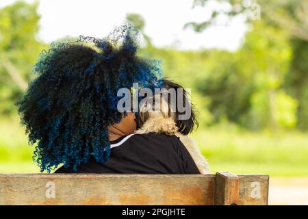 Goiania, Goias, Brasilien – 20. März 2023: Foto einer jungen Frau von hinten, die mit einem Hündchen auf einer Bank sitzt. Stockfoto