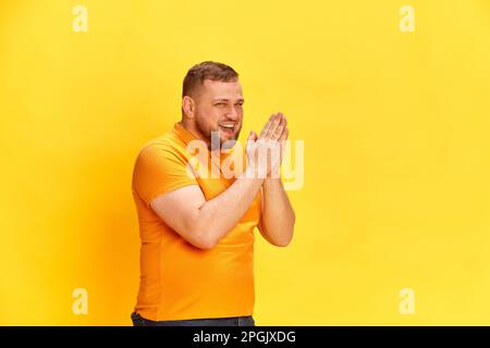 Porträt eines lachenden Mannes mit lächelndem Gesicht, der steht und Hände über gelben Hintergrund reibt. Schließen Stockfoto