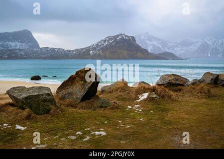 Steine auf Gras, Strand Haukland und Nordatlantik, felsige Berge bei windigem Wetter am Ende des Winters. Lofoten-Inseln, Norwegen. Stockfoto