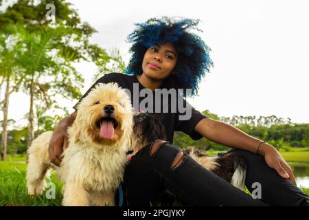 Goiania, Goias, Brasilien – 20. März 2023: Eine junge schwarze Frau mit lockigem Haar, blau gefärbt, die mit ihren Hunden auf dem Rasen eines Parks sitzt. Stockfoto