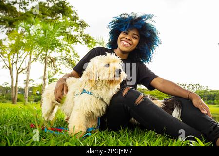 Goiania, Goias, Brasilien – 20. März 2023: Eine junge schwarze Frau mit lockigem Haar, blau gefärbt, die mit ihren Hunden auf dem Rasen eines Parks sitzt. Stockfoto