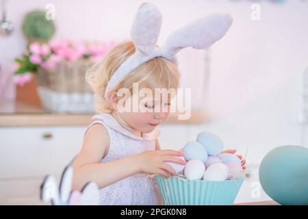 Porträt eines süßen kleinen Mädchens in Osterhäschenohren legt bemalte Eier auf einem blauen Teller in der hellen Küche zu Hause. Stockfoto