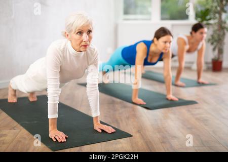 Konzentrierte, reife Frau, die in der Dolphin-Plank-Pose oder Makara Adho Mukha Asana während des Gruppen-Yoga-Kurses im Studio steht Stockfoto