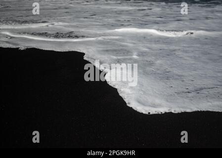 Meist verschwommener schwarzer Sandstrand-Hintergrund mit weißem Schaum von Meereswellen Stockfoto