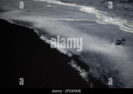 Meist verschwommener schwarzer Sandstrand-Hintergrund mit weißem Schaum von Meereswellen Stockfoto