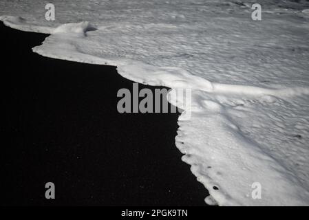 Meist verschwommener schwarzer Sandstrand-Hintergrund mit weißem Schaum von Meereswellen Stockfoto