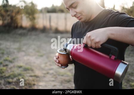 Junger lateiner, der Yerba-Mate trinkt, heißes Wasser aus einer roten Thermoskanne serviert, auf dem Land. Stockfoto