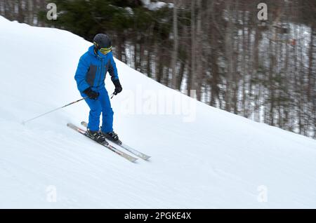 Ein Mann in blauer Sportbekleidung auf Skiern fährt einen schneebedeckten Abhang hinunter Stockfoto