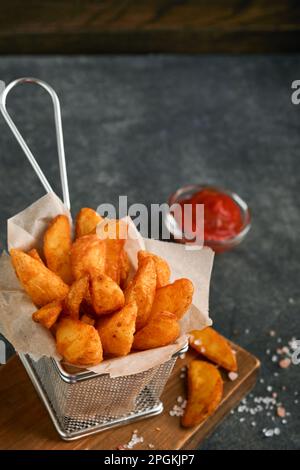 Pommes frites in einem Metalldrahtkorb mit Salz und Ketchup auf altem dunklem Holzkloake. Bratkartoffeln. Fast Food und ungesunde Nahrungsmittel Konzept. Stockfoto