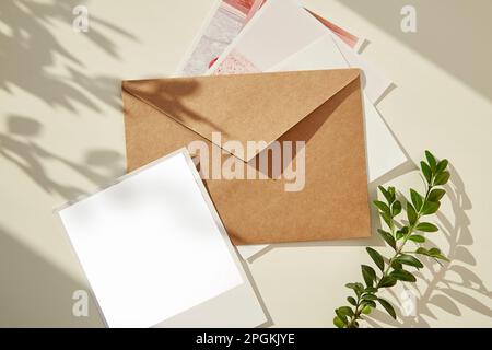 Beigefarbener Papierumschlag mit Fotografiemodell, grüner Zweig unter Schatten. Sammlung von Grußkarten, Postkarten, Einladungen. Flach verlegt. Stockfoto