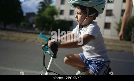 Das Kind lernt mit seiner Mutter, draußen mit einem Helm zu fahren. Eltern, die kleinen Jungen Fahrrad beibringen. Kindliche Entwicklung Stockfoto