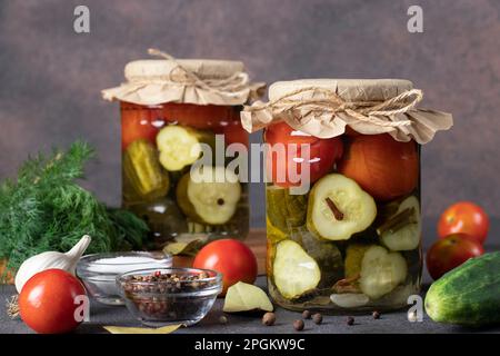 Eingelegte Gurken mit Kirschtomaten in zwei Glasgefäßen auf braunem Hintergrund mit frischen Zutaten Stockfoto