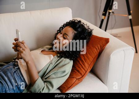 Fröhliche junge afrikanische Frau, die ihr Smartphone benutzt, während sie auf dem Sofa in einer gemütlichen Wohnung liegt Stockfoto