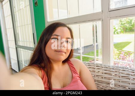 Nahaufnahme des Selfie mit dem Mobiltelefon, Porträt einer jungen Frau, die lächelt und in die Kamera schaut, zu Hause sitzt und ein Bild von sich selbst macht Stockfoto