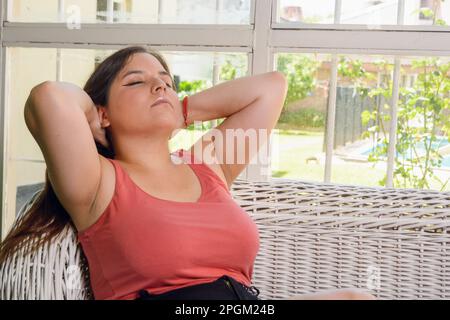 Eine junge lateinin lehnt sich zu Hause auf die Couch, mit den Händen hinter dem Hals und geschlossenen Augen, ruht sich aus, ist ruhig und denkt nach Stockfoto